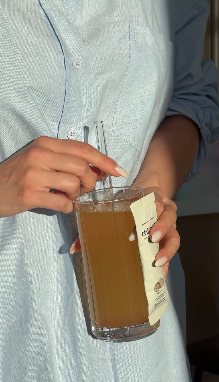 A person in a light blue button-up shirt holds a tall clear glass filled with a brown beverage and a clear straw, with a cream-colored product packet attached to the side.